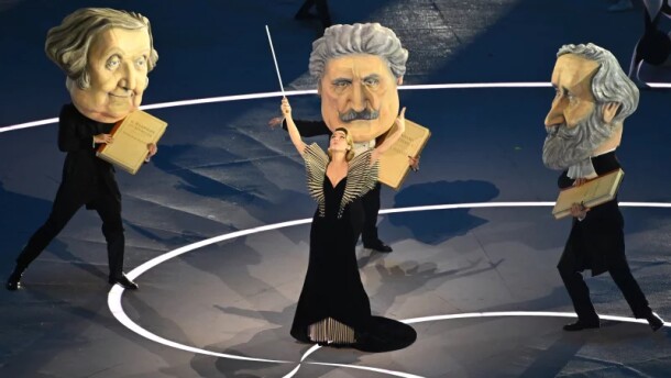 A woman in a black dress conducts three dancers dressed as famous Italian composers: Rossini, Puccini, and Verdi.