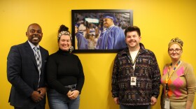 Four people stand in front of a yellow wall with a npicture of a graduating student