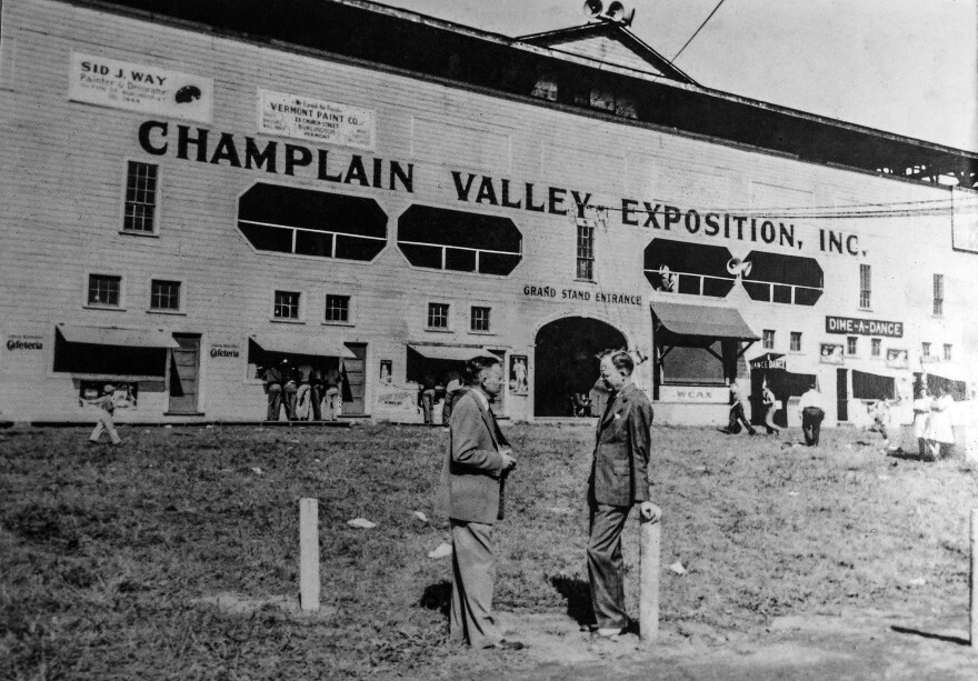 Two men in suits stand in the forefront of an old black-and-white photo. A large building in the background with the words, "Champlain Valley Exposition, Inc.," is painted on its front. Others are gathered in small groups at small vendor stands.