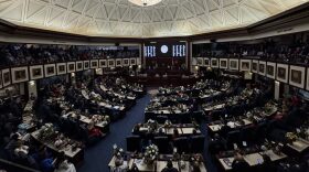 Lawmakers gathered in the Florida House of Representative chambers. A desk in the middle as other tables circle it in a round room with a dome ceiling. blue carpeting, white and brown paneling