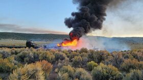 Side view of the remains of a semi-truck on a remote road, which is charred and nearly unrecognizable. There's a large flame and a huge plume of black smoke.