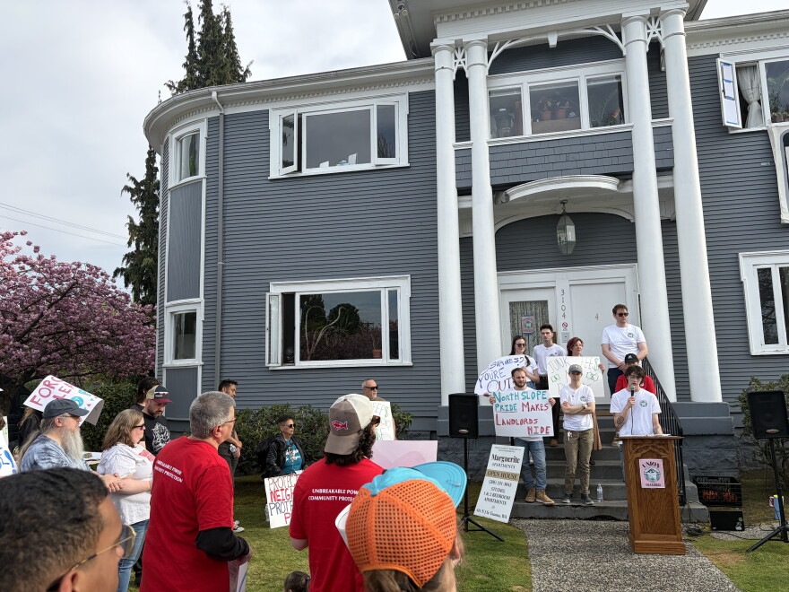 People speak outside of a small apartment complex in Tacoma. They hold signs that say "Union strong: and "North Slope Pride makes Landlords hide" 