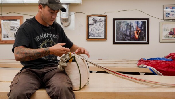 A man sits on a table and packs a parachute. There is a picture of a wildfire on the wall behind him.