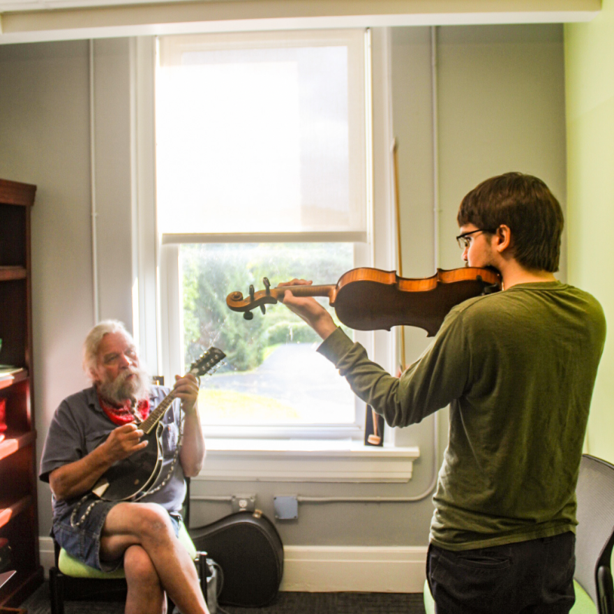 A person is playing a violin while another person plays banjo in front of a window.