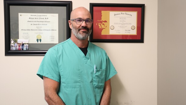 Dr. Michael Fenech stands in front of a wall in his office. He's wearing teal colored scrubs. The wall has two framed diplomas.