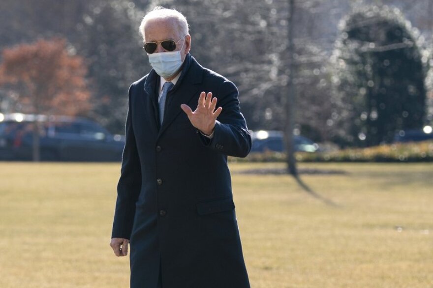 President Joe Biden, wearing a dark blue long coat, sunglasses and a face mask, waves to reporters as he arrives on the South Lawn of the White House.