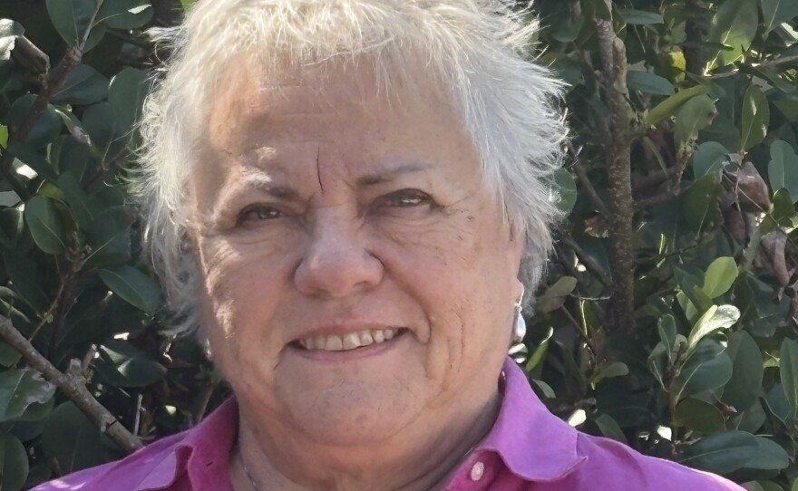 A smiling woman with short white hair wearing a purple button-down shirt and silver necklace
