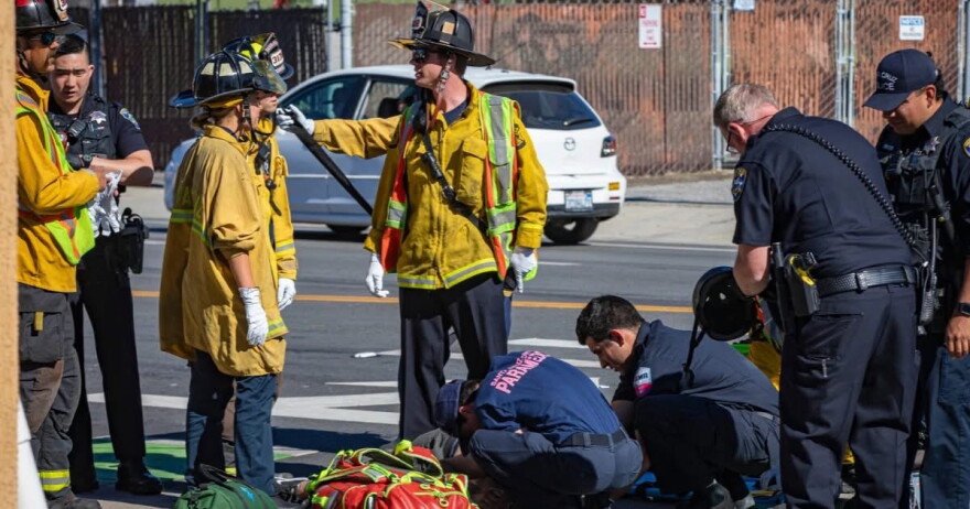 Firefighters in yellow jackets stand on the left while paramedics in blue attend to a person lying on the ground.