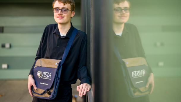 Austin Draper stands next to a reflective wall wearing a blue and brown bag with the VISTA logo.
