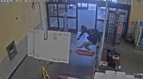A ceiling mounted camera catches a man jumping over a turnstile inside a store. The back of a flatscreen TV is in the foreground.