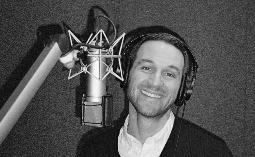 A black and white photo of a man with short dark hair, mustache, and beard, wearing headphones and standing in front of a microphone. He is wearing a white button-down shirt under a dark sweater.