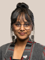 An Indian person with hair in a bun, chunky black earrings and a grey, red and black blazer.