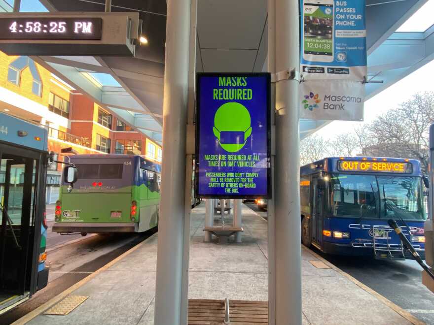 A walkway with busses on either side of it, with a digital poster in the middle describing that masks are required to board the bus.