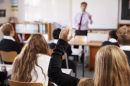 Un estudiante levanta la mano en un aula, junto a otros niños y con el profesor al frente.