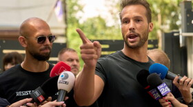 A blad white man wearing sunglasses on the left as his brother, a white man with a receding hairline points a finger outward as he’s surrounded by news microphones.