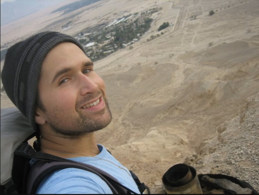 Lee Frankel-Goldwater, a man wearing a light blue T-shirt and stocking cap, with a backpack, looks over his shoulder with trees and buildings in the distance
