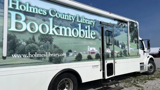 The Holmes County Library Bookmobile is parked in a gravel lot. It's a white truck covered in a photo of rolling hills with a red farm house.