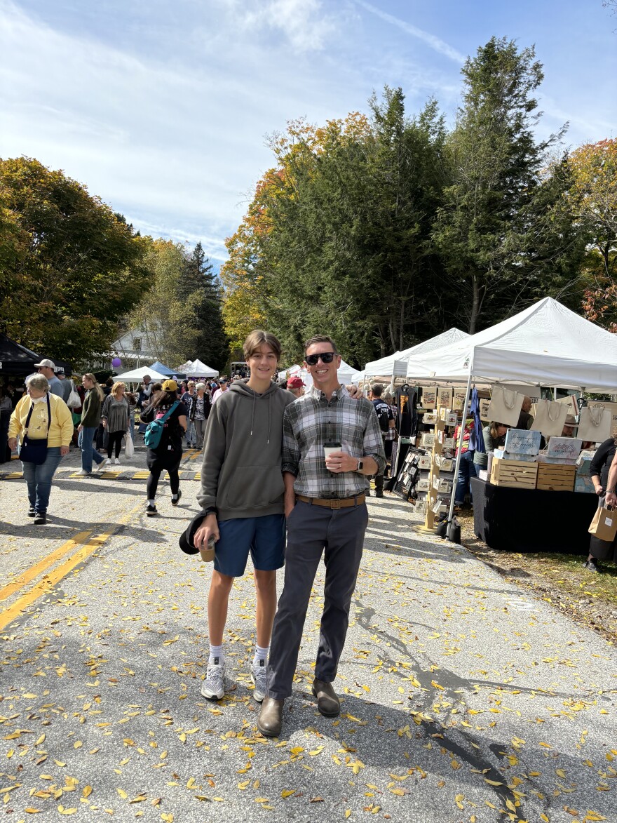 A young man stands next to his father at an outdoor fall market.