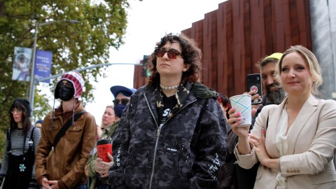 Under a cloudy sky, several people are looking towards someone speaking in front of them. They are wearing a variety of clothes. 