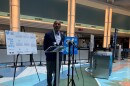 Stewart speaking at a podium at the entrance of the airport terminal, with a bunch of signs around him about COVID-19 guidelines and more airport precautions.