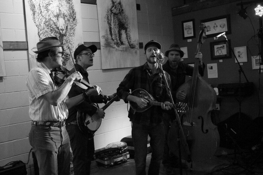 Four men gathered around a microphone playing fiddle, banjo, mandolin, and stand up bass and singing