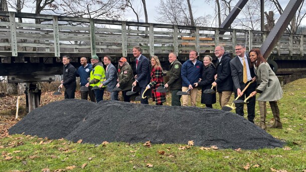 Gov. Glenn Youngkin (center), with 12 other federal, state and local officials, gather around a mound of grey crushed gravel, the same material used in the Virginia Creeper Trail. They hold shiny shovels, aimed at the mound. Behind them is a wooden bridge from the trail. 