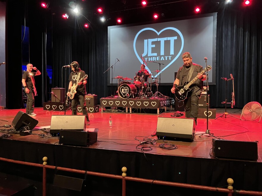 A band, all in black, is on a stage washed red playing their guitars and drums. In the background, an image says Jett Threatt. 