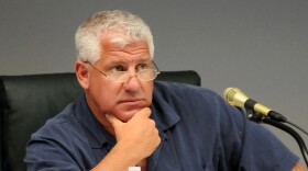 Man with white hair puts hand on chin sitting next to mic