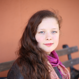 A woman with long, curly, brown hair and an eyebrow piercing wears a pink scarf and a black coat. She smiles at the camera, facing forward but turning to the side
