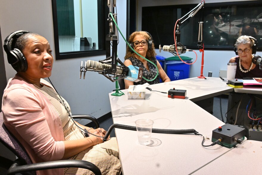 Three women sit around a table in a radio studio. Each is near a microphone. The woman at left is talking. The others are looking at her and listening.