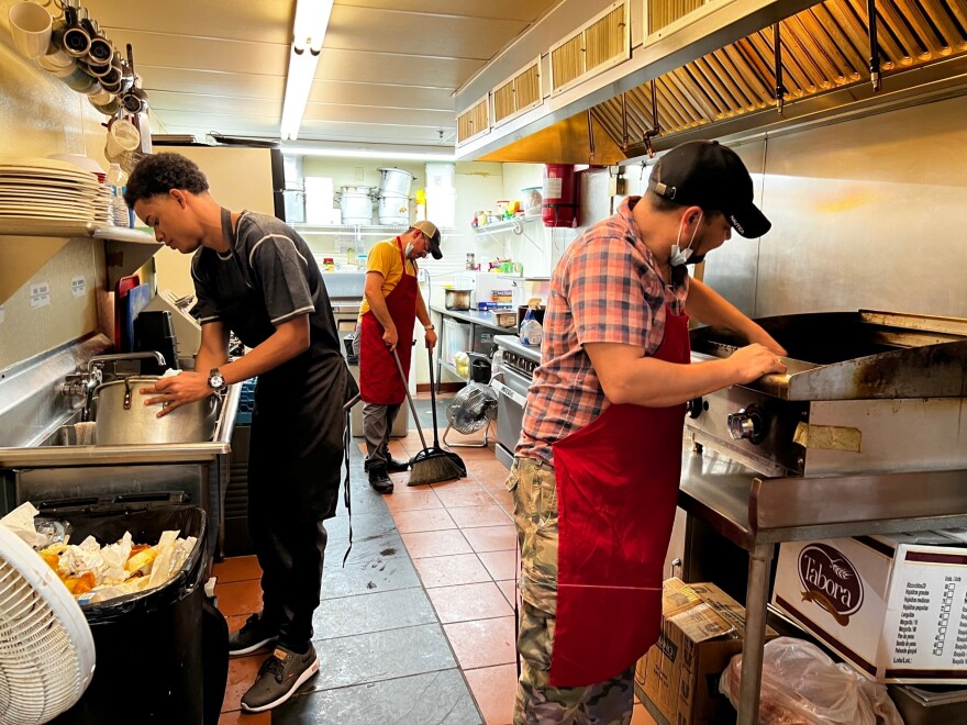 Three men work inside a restaurant kitchen. One is washing dishes, one is sweeping the floor and the other is cleaning a cooking grill.