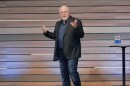Finance guru Dave Ramsey wears a black blazer and jeans and talks on a stage in front of a wood paneled background