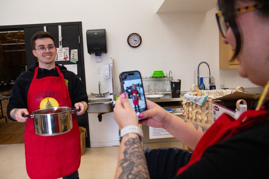 A photo of a young person holding up a phone to photograph another young person holding a pot and wearing an apron that says wlipogwat in a kitchen