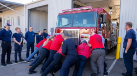 A group of around 9 people push a fire engine into its bay. Most of them are wearing red shirts. They are surrounded by onlookers wearing navy shirts. A man sits in the driver's seat of the fire engine. 