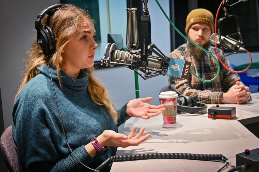 Two people sit in a radio studio. The woman at left is talking at a microphone and gesturing with two hands. The man, sitting at right, is listening.