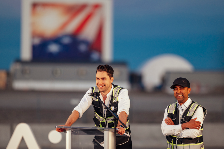 A man in a hi-visibility vest stands at a microphone beaming while another man in a vest smiles beside him.