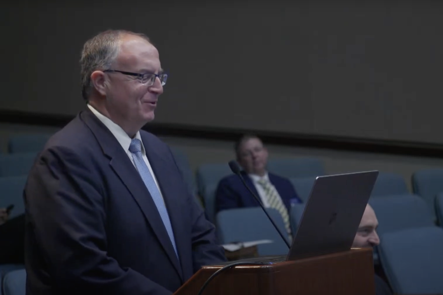 John Keller stands at a podium and speaks into a microphone. He wears a blue suit and reads off of a laptop.