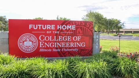 Large red sign that reads 'Future Home of the College of Engineering at Illinois State University' in front of a large office building and behind several plants. 