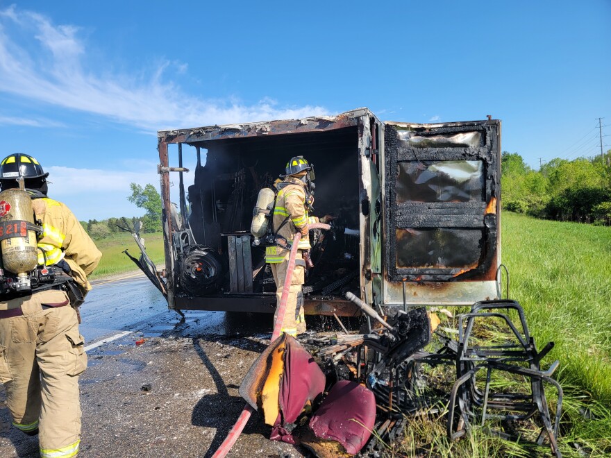 Two firefighters stand with hoses, extinguishing  the remnants of a fire from near a charred van on rural higway.