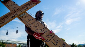 A man carrying a wooden cross also wears a mask, gloves and glasses during a march near Music Hall as people continue to rally and protest the murder of George Floyd as the coronavirus pandemic continues, Wednesday, June 3, 2020, in Cincinnati.