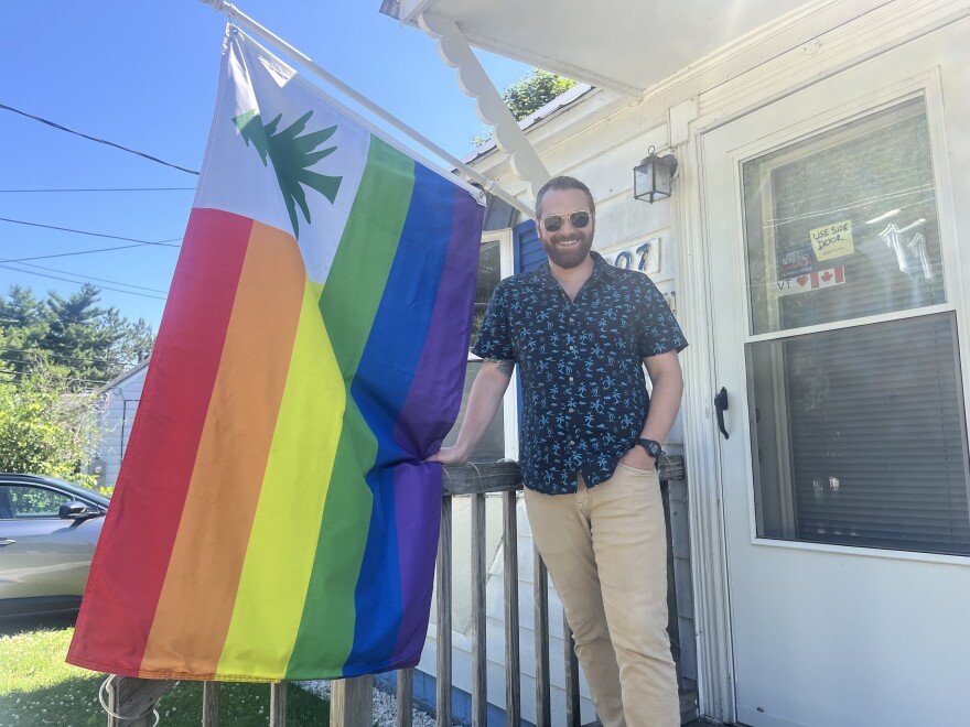 A man in a patterned shirt stands in front of a rainbow striped flag with a tree in the corner.