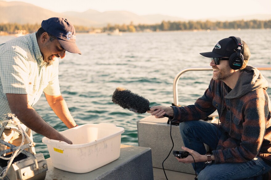 A person standing on a boat and leaning over a plastic bin. They are being interviewed by another person who is wearing headphones while sitting and holding a microphone toward them.