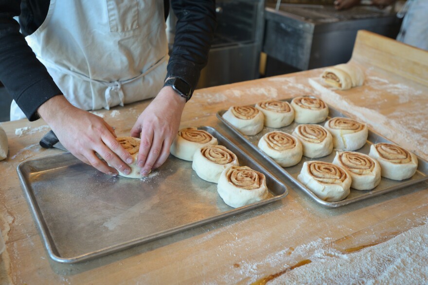 A pair of hands sets down one unbaked cinnamon roll into one of two tray of cinnamon rolls that has three other unbaked rolls. Next to that pan is a baking pan filled with nine unbaked cinnamon rolls.