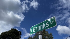 A street sign that reads "Brunks Lane" stands in front of a tree line and a blue sky with puffy white clouds.