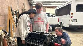 Two students stand over a car engine while a man explains the mechanics.