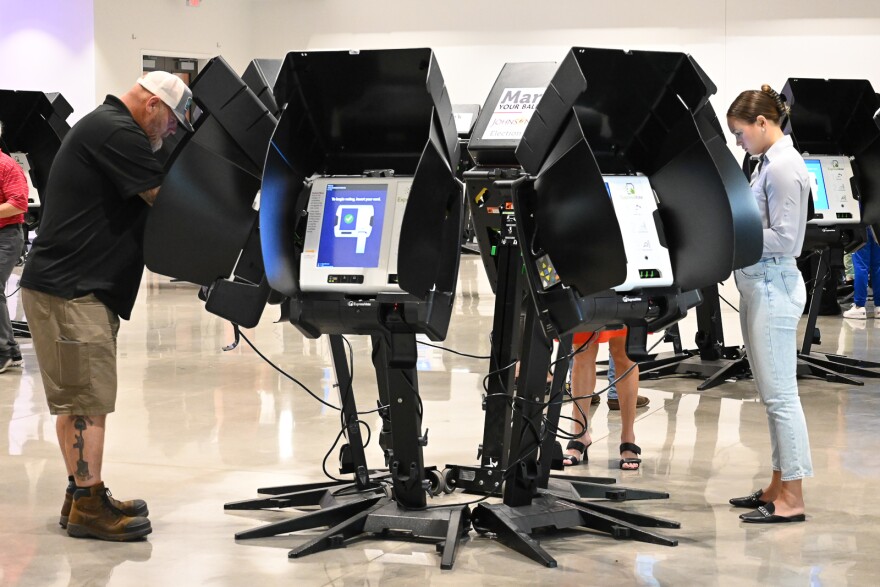 A man and a woman standing on either side of a cluster of electronic voting machines cast their votes.