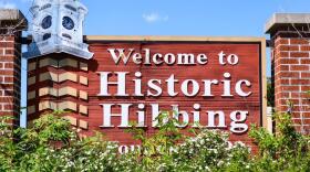 A wooden sign with a red hue between two brick pillars reads, "Welcome to Historic Hibbing." The sign has a clock tower on the left-hand side. The bottom of the sign is covered by a green bush with small white flowers.
