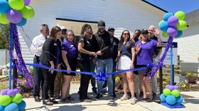A group surrounds two people who are using a giant pair of scissors to cut the ribbon leading to the white building behind them 