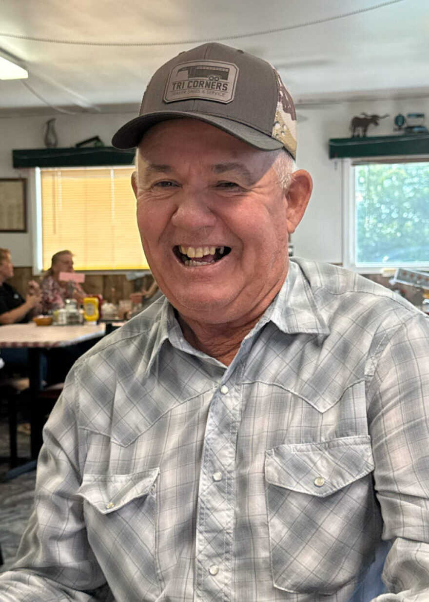 An older white man wearing a gray pearl snap button down and a trucker cap that says, "Tri corners."
