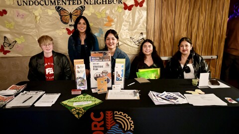 Four volunteers sitting at a table with resource pamphlets for struggling families and people who want to get involved.  Jaqueline Vazquez is standing behind them and there's a poster on the wall with the words "Love thy undocumented neighbor" and butterflies of various colors.